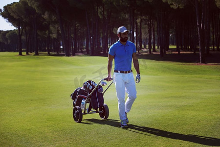 男人走路摄影照片_在阳光明媚的日子里，帅气的中东高尔夫球手背着轮包在球场上散步。高尔夫球手背着轮包走路