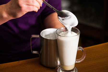 咖啡倒了摄影照片_咖啡师正在往杯子里倒牛奶泡沫，里面有焦糖--准备了又甜又辣的咖啡拿铁。咖啡师正在准备拿铁咖啡