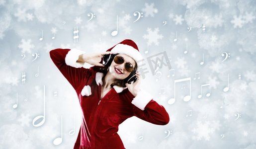 音乐圣诞老人摄影照片_庆祝新年。年轻漂亮的圣诞老人女孩戴着耳机听音乐