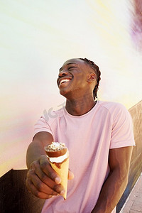 黑色t恤摄影照片_年轻英俊的黑人男子穿着一件粉色的t恤，微笑着拿着一个冰激凌蛋筒，在夏天，在一面画着的墙上，像是日出或晴朗的日子