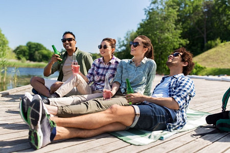 喝啤酒摄影照片_休闲、野餐和人的概念-朋友们在夏季公园的湖畔码头喝啤酒和苹果酒。朋友们在湖边码头喝着啤酒和苹果酒