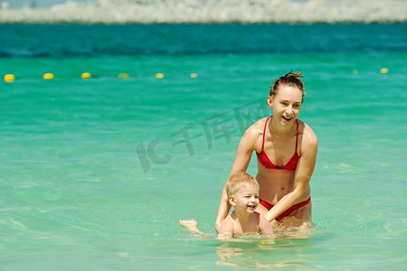 教游泳摄影照片_快乐的妈妈教她的小儿子游泳。两岁的小男孩和妈妈一起在海滩上。快乐的妈妈教她的小儿子游泳。 