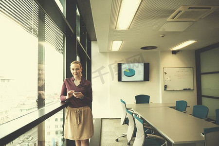 商务潮人摄影照片_在现代创业公司商务办公室会议室内部，年轻的女商人穿着休闲的时髦服装，使用智能手机