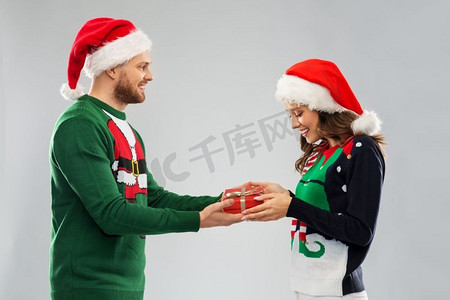 圣诞毛衣摄影照片_人们和节日的概念-在丑陋的毛线派对上，戴着圣诞帽的幸福夫妇带着圣诞礼物。穿着圣诞毛衣和礼品盒的幸福夫妇
