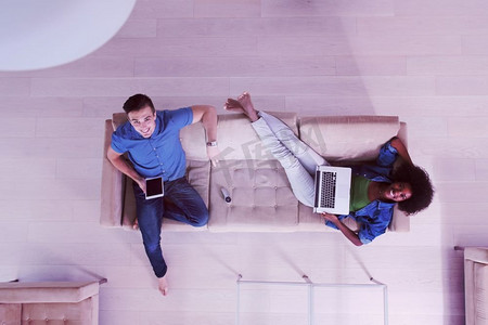 年轻的多民族夫妇在豪华客厅里放松，使用平板电脑和笔记本电脑俯视