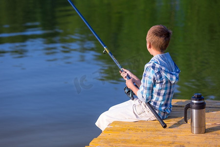 穿着蓝色衬衫的男孩坐在海边的码头上，手里拿着钓鱼竿