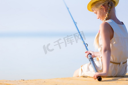 女孩穿着裙子，戴着一顶带钓鱼竿的帽子。穿着连衣裙戴着帽子的女孩在码头钓鱼