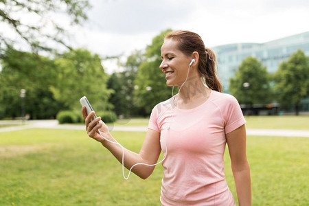 健身、运动和健康生活方式理念-公园里微笑的女人在智能手机上听音乐。一名女子在公园里用智能手机听音乐