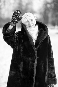 一个微笑的高级成年妇女戴着羊毛帽，围巾和彩色手套，用她的手敬礼的冬季肖像，与雪背景