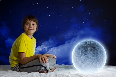 晚上做梦。可爱的男孩坐在床上与月球行星