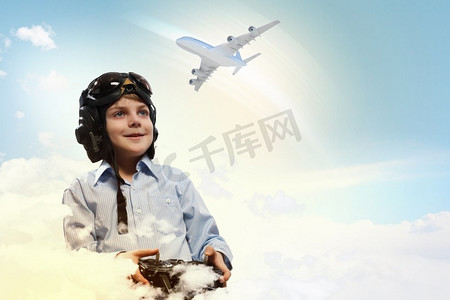 玩玩具的男孩摄影照片_戴着飞行员头盔的小男孩在云的背景下玩玩具无线电飞机