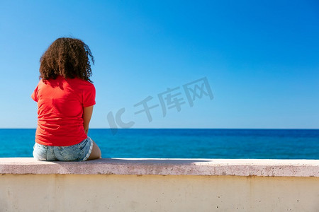 混血黑人少女女青年身穿红色T恤和牛仔短裤坐在墙上眺望蔚蓝大海