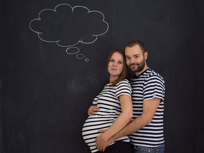 丈夫和怀孕的妻子在黑色粉笔画板前摆姿势的肖像。怀孕夫妇在黑色粉笔画板前摆姿势