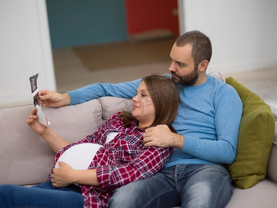 怀孕的夫妇看起来像婴儿&S；超声波。坐在家里的沙发上休息时，年轻的怀孕夫妇看起来像婴儿和S的超音波照片