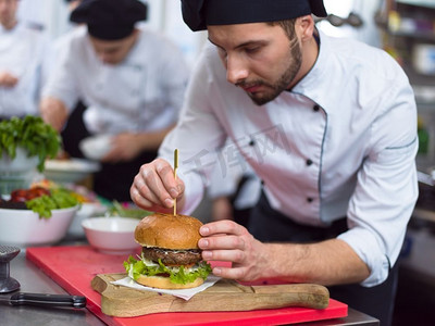 快餐厅摄影照片_厨师大师在餐厅厨房的汉堡包上放牙签。厨师精选汉堡