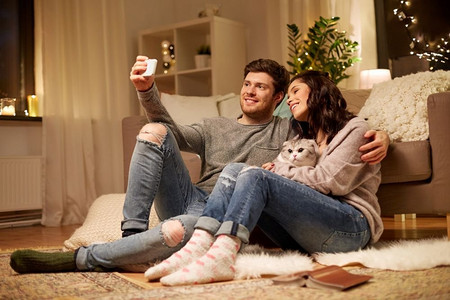休闲、技术和人概念-与猫的愉快的夫妇采取通过智能手机的selfie在家快乐的夫妇采取selfie通过智能手机在家里