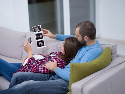 怀孕的夫妇寻找婴儿的超声波。’年轻的怀孕夫妇寻找婴儿的超声波照片，而放松在沙发上在家里’