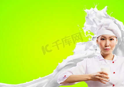 亚洲女厨师防止牛奶溅起。亚洲女厨师手持一杯牛奶，穿着红色围裙，手持牛奶，在彩色背景下飞溅