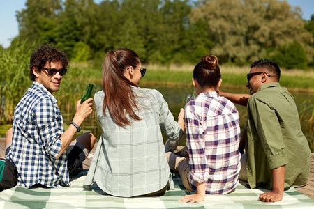 休闲，野餐和人的概念—朋友喝啤酒和苹果酒在湖码头在夏季公园。朋友们在湖码头喝啤酒和苹果酒