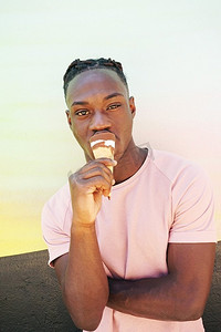 年轻英俊的黑人男子穿着一件粉色t恤，手持冰激凌蛋卷，在阳光明媚的夏天，在一面画好的墙上吃着。