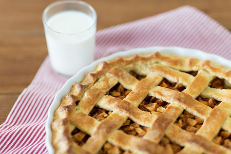 一杯牛奶杯摄影照片_食物、烹饪和烘焙概念--在木桌上放苹果派、一杯牛奶和厨房毛巾。烘焙模具中的苹果派和一杯牛奶