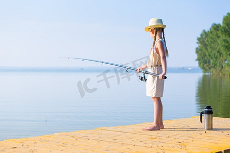 带帽子的女孩摄影照片_女孩穿着裙子，戴着一顶带钓鱼竿的帽子。穿着连衣裙戴着帽子的女孩在码头钓鱼