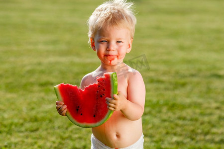 蹒跚学步的孩子的肖像户外。农村场景与一岁的男婴吃西瓜片在花园里。一张脏兮兮的快乐的脸。