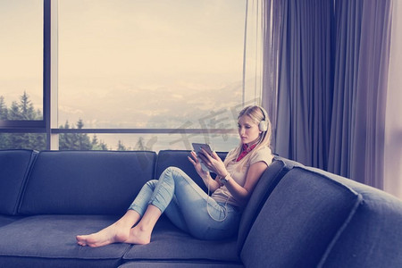 年轻漂亮的女孩戴着耳机享受音乐，躺在家里靠窗的沙发上。戴着耳机欣赏音乐的年轻女孩