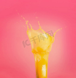 粉色夏天背景摄影照片_带有黄色溅起的夏季饮料的玻璃杯：粉红色背景下的奶昔或果汁，正视