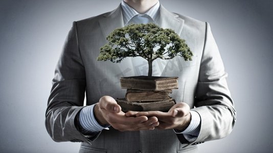 开发你的智力。男人拿着一堆书，手里拿着一棵正在生长的绿树的特写