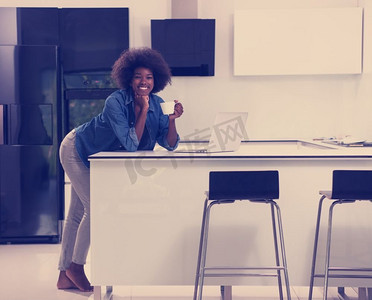 年轻面带微笑的黑人妇女在现代厨房里一边玩电脑，一边喝咖啡