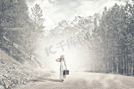 她轻装上阵。穿着白色长裙，戴着帽子的行李箱在森林路上的女人