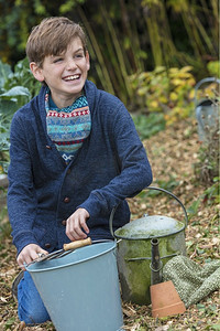 花园菜地摄影照片_快乐微笑的男童在菜地里用水桶、花园叉子和水壶园艺