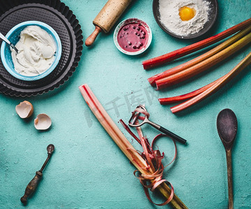 果皮剥落。  新鲜的红色大黄茎与剥皮器在厨房桌子背景有烹调和烘焙工具，顶视图季节性食物概念