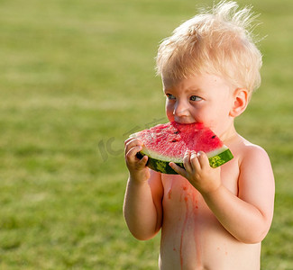 吃西瓜场景摄影照片_户外蹒跚学步的孩子的肖像。一岁的男婴在花园里吃西瓜片的农村场景。快乐的孩子肮脏的脸。