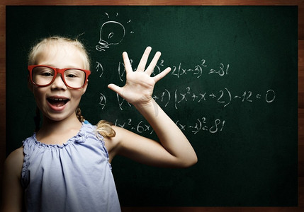 儿童数学摄影照片_聪明的女学生。戴着红眼镜的天才女孩在黑板上写着公式