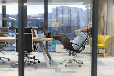 这是放松的时候。疲惫的年轻休闲商人在办公室的办公桌前放松