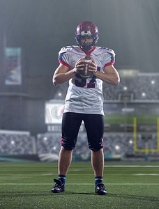 自信的美国橄榄球运动员的画像拿着球当站立在大的现代体育场与灯和照明弹在晚上