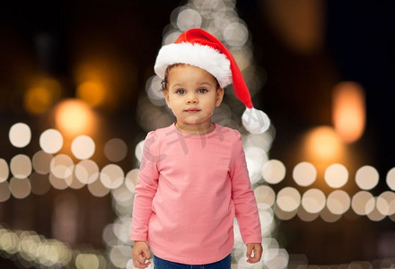 节日和人们的概念-圣诞帽上的小女婴圣诞树上的灯光背景。圣诞节戴着圣诞帽的小女婴