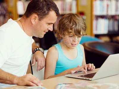 带学生摄影照片_小男孩和他的父亲在图书馆里带着笔记本电脑。小男孩和他的父亲带着笔记本电脑