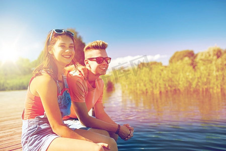 假日、假期、爱情和人的概念--夏天坐在河边泊位上的幸福的青少年情侣。幸福的青少年夫妇坐在河边的泊位上
