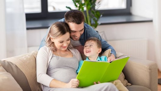 怀孕，人和家庭观念-快乐的怀孕母亲，父亲和小儿子在家里的沙发上看书。快乐的家庭在家读书