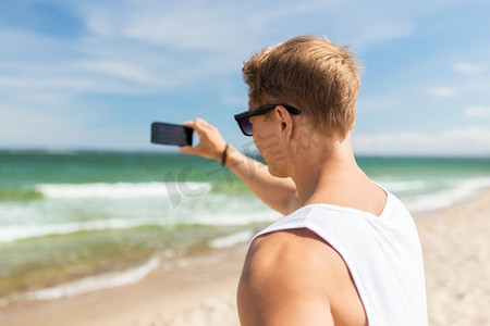 暑假和人的概念-年轻人在海滩上用智能手机拍摄大海。一名男子在夏日海滩用智能手机拍照
