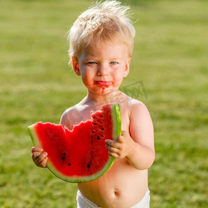 吃西瓜场景摄影照片_户外学步儿童的肖像。一岁男婴在花园里吃西瓜片的乡间场景。快乐孩子脏兮兮的脸。