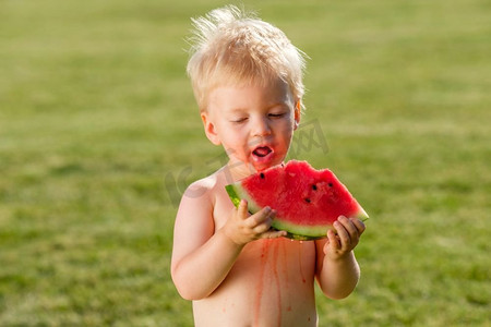 蹒跚学步的孩子的肖像户外。农村场景与一岁的男婴吃西瓜片在花园里。一张脏兮兮的快乐的脸。