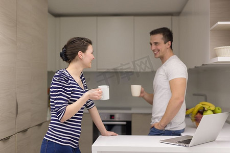 幸福的年轻夫妇手持笔记本电脑，在家中的现代厨房里享用早餐咖啡