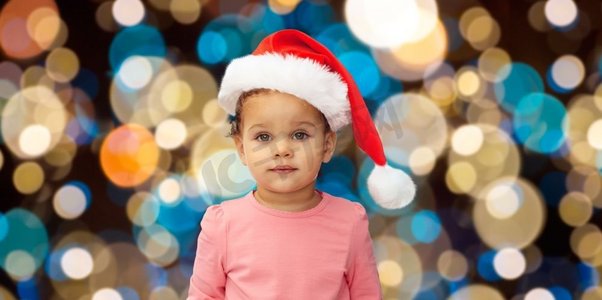 圣诞节，节日和人们的概念-戴着圣诞帽的小女孩在灯光下的背景。圣诞节戴着圣诞帽的小女婴