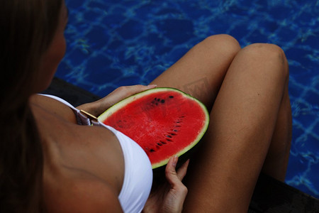 在泳池边拿着西瓜的女孩。女孩抱着西瓜在蓝色的池塘里，修长的腿。热带水果饮食。夏日田园诗般的假期。