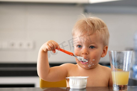吃酸奶摄影照片_可爱的一岁男孩用勺子吃酸奶。肮脏凌乱的脸蹒跚学步的孩子。