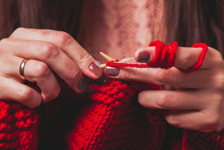 织毛衣的针摄影照片_手握着针，编织着一件红色的保暖夹克。女性手织毛衣
